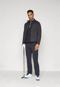 Mees mustas golfiülikonnas, hoides putterit, kandes valget golfikinda ja valgeid golfijalatseid siniste talladega, seismas ühe värvilise tausta ees.