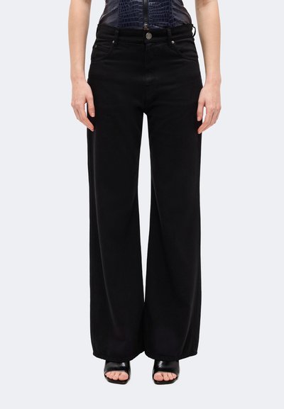 Modèle portant un jean noir taille haute à jambes larges et droites, ainsi que des sandales noires à bout ouvert.