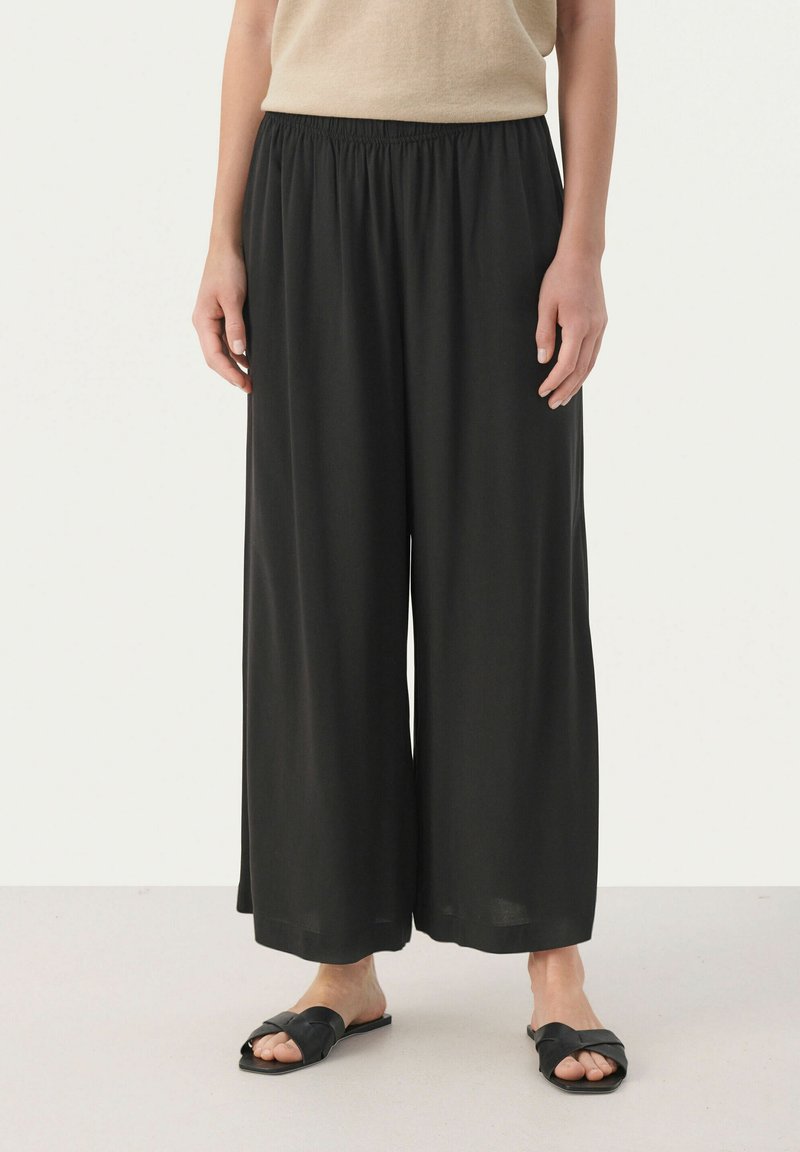 Pantalons larges noirs avec une taille élastique, fabriqués en tissu doux. Portés avec des sandales plates noires, présentant une coupe décontractée.