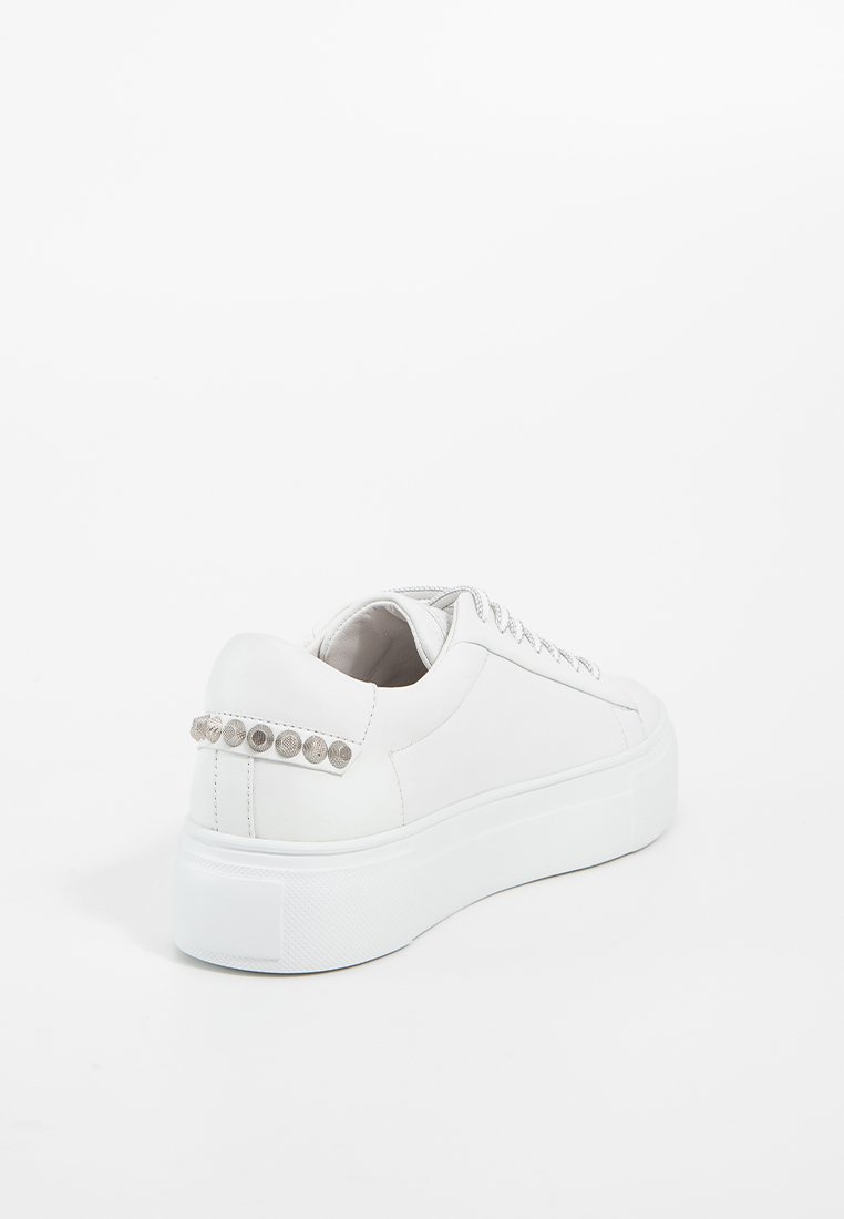 Weiße Low-Top-Sneaker mit dickem Sohlenrand und metallischen kreisförmigen Nieten an der hinteren Fersenschlaufe, fotografiert vor einem weißen Hintergrund.