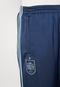 Jūros karinių atletinių kelnių su šviesiai mėlynomis šoninėmis juostomis, užtrauktuku užsegama kišene ir Ispanijos herbu, kuriame yra karūna ir žvaigždė.