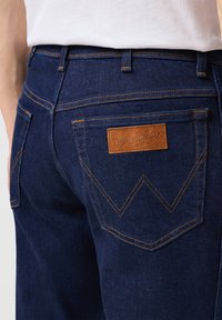 Jeans de mezclilla azul oscuro con un acabado texturizado, que presentan costuras naranjas y un parche de cuero marrón en el bolsillo trasero.