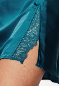 Indumento in raso turchese con bordo in pizzo smerlato lungo la manica, con una texture liscia e un design a vestibilità rilassata.