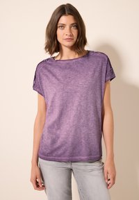 T-shirt violet ample avec un col en scoop, des manches courtes et des accents de boutons sur les épaules ; fabriqué dans un tissu doux et texturé.