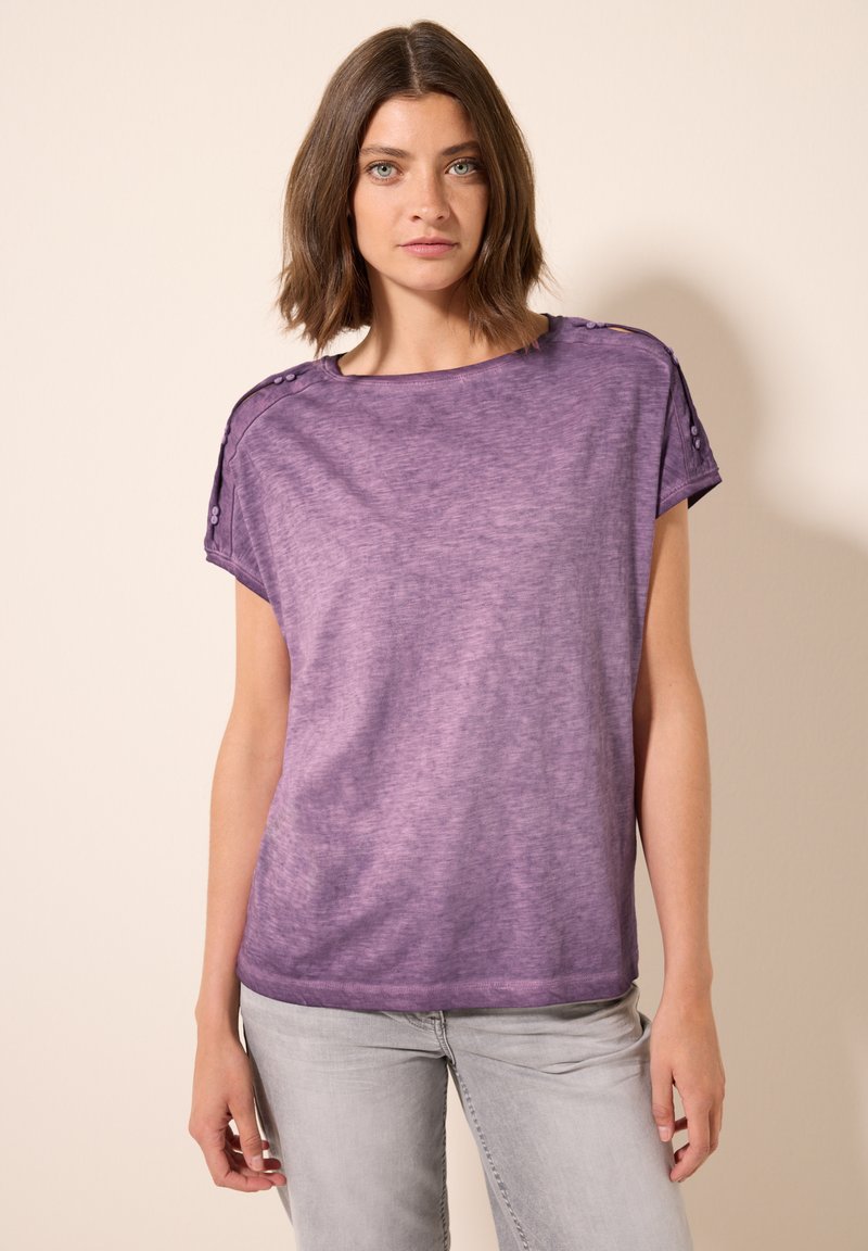 T-shirt violet ample avec un col en scoop, des manches courtes et des accents de boutons sur les épaules ; fabriqué dans un tissu doux et texturé.