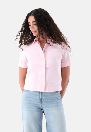 Frau mit lockigem Haar, die ein hellrosa kurzärmeliges Hemd mit Knöpfen und hellblaue hochgeschnittene Jeans trägt und nach unten und zur Seite schaut.