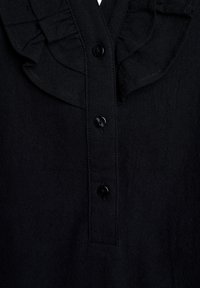 Schwarzes Hemd mit Rüschensaum, ausgestattet mit einer durchgehenden Knopfleiste und strukturierter Stoffqualität. Sechs schwarze Knöpfe entlang der Knopfleiste.