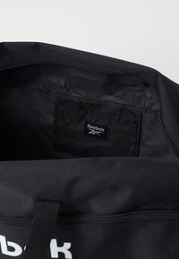 Μαύρη αθλητική τσάντα με φερμουάρ στην κορυφή, διαθέτει μια μικρή εσωτερική τσέπη και εμφανές λογότυπο της Reebok στο μπροστινό μέρος. Λείο υφαντό υλικό.