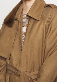 Brązowy trencz z wyprofilowanym kołnierzem, paskiem w talii i gładką teksturą. Pod spodem częściowo widoczny jest sweter w wzory.