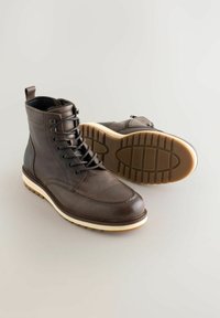 Un par de botas de cuero marrón con cordones y suelas de goma color crema, una bota erguida y la otra mostrando la suela texturizada.
