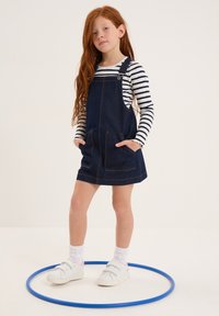 Abito in denim blu scuro con bretelle, dotato di due tasche frontali, abbinato a una maglietta a righe bianche e nere a maniche lunghe. Scarpe bianche, hula hoop blu.