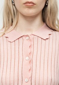 Κοντινή λήψη ενός ατόμου που φοράει ροζ πλεκτό τοπ με κυματιστό γιακά και κάθετες ανοικτές πλεκτές ρίγες, κουμπωμένο με λευκά κουμπιά.