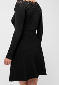 Robe noire côtelée à manches longues, détail en dentelle dénudée des épaules, taille cintrée et jupe évasée. Tissu texturé avec un motif subtil.