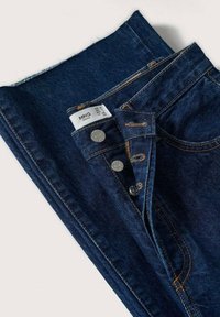 Jean denim bleu foncé plié avec fermeture par boutons et étiquette de taille blanche visible à l'intérieur de la ceinture.