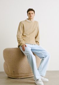 Beige ribbed knit sweater, light blue jeans, and white sneakers, seated on a rounded, textured beige upholstered stool against a neutral backdrop.
