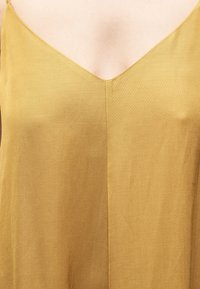 Γυναίκα που φοράει μουσταρδί κίτρινη αμάνικη μπλούζα με V-λαιμόκοψη και εμφανές κεντρικό ραφάκι μπροστά.