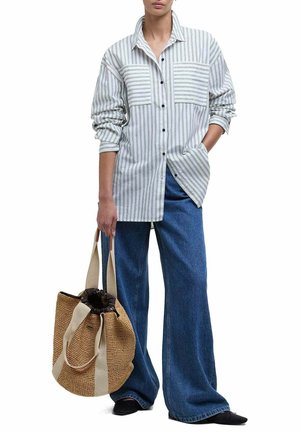 Vrouw die een oversized gestreept overhemd met knopen en wijde blauwe jeans draagt, en een grote gevlochten totebag met beige banden vasthoudt.