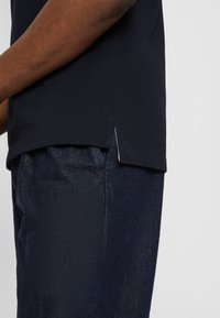 T-shirt in cotone blu scuro con orlo dritto e dettagli con spacco laterale, abbinato a jeans in denim scuro con una leggera texture.