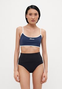 Marineblå crop top med hvit kant og logo; kombinert med høy midje, svarte bikinibukser. Glatt stoff, minimal design, tettsittende form.