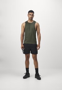 Top sin mangas de color verde oscuro, pantalones cortos negros, calcetines negros y zapatillas deportivas. El modelo sostiene equipo de entrenamiento con un fondo neutro.