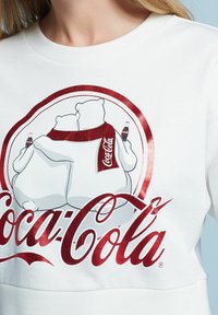 Sweatshirt blanc présentant un graphique de deux ours polaires s'étreignant, avec un branding Coca-Cola en rouge et blanc, soulignant le design ludique.