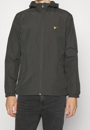 Schwarze Kapuzenjacke mit Reißverschluss, elastischen Bündchen und kleinem gelbem Vogel-Logo auf der linken Brust, getragen von einer Person mit schwarzer Jeans.