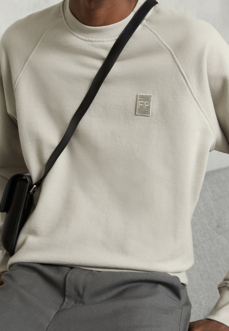 Personne portant un sweat-shirt beige clair avec un patch logo FP, un pantalon gris et un sac bandoulière noir reposant sur leurs genoux.