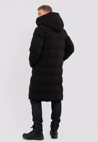 Lungo cappotto imbottito nero con cappuccio, caratterizzato da sezioni trapuntate orizzontali, indossato sopra jeans scuri e stivali neri.