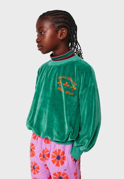 Grüner Samt-Sweatshirt mit rundem Ausschnitt, gestickten orangen Blumen und langen Ärmeln. Dazu pinkfarbene Hosen mit floralem Muster.