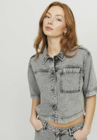 Chaqueta de mezclilla gris de manga corta con corte corto, cierre de botones y un bolsillo en el pecho; cuenta con costuras en contraste y un acabado texturizado.