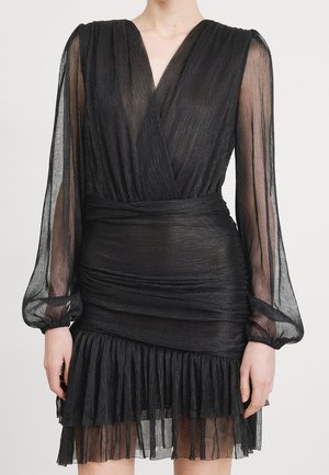 Femme portant une mini-robe plissée noire avec des manches longues transparentes, un décolleté en V profond et un ourlet à volants superposés.