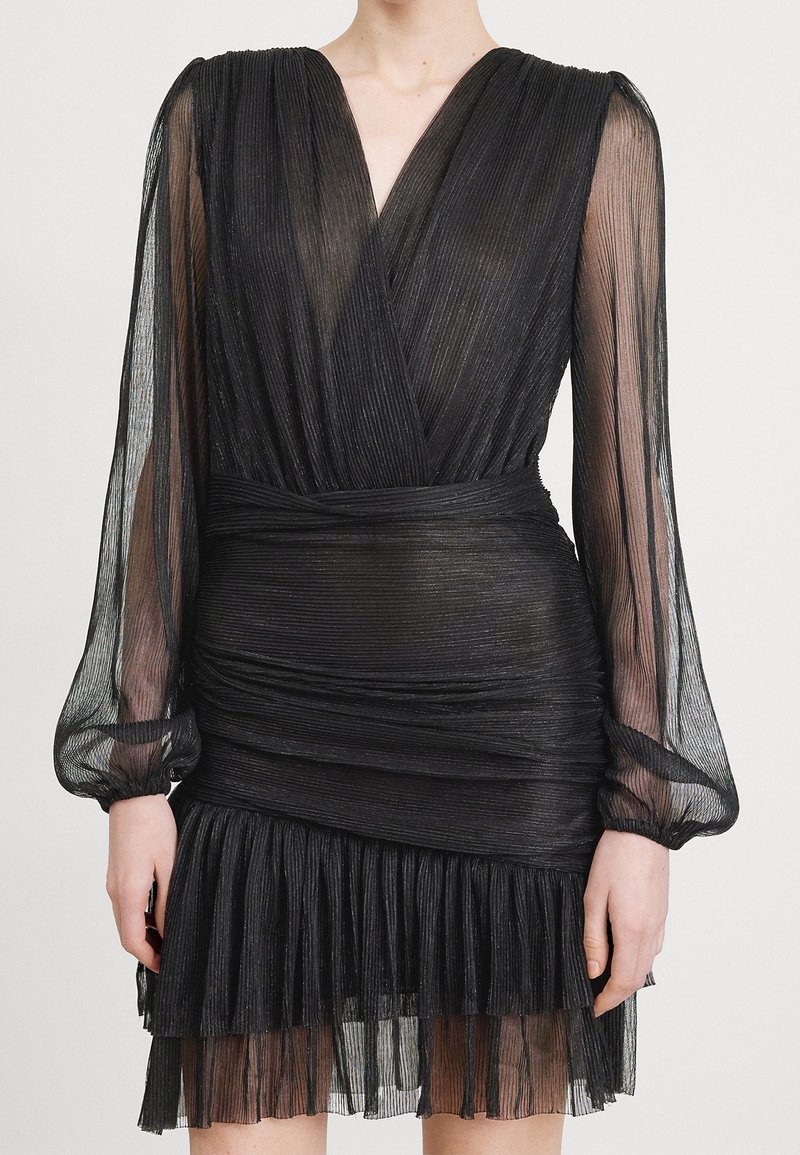 Femme portant une mini-robe plissée noire avec des manches longues transparentes, un décolleté en V profond et un ourlet à volants superposés.