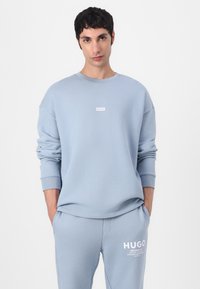 Lichtblauwe sweatshirt en bijpassende joggingsbroek met "HUGO" logo. De stof heeft een soepele textuur, ontworpen met een oversized pasvorm en geribde manchetten.