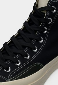 Μαύρο πάνινο ψηλό παπούτσι με λευκή ραφή, μεταλλικά οπές και μαύρα κορδόνια, με αντίθεση από μπεζ καουτσούκ σόλα.