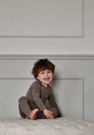 Barna, hosszú ujjú body kerek nyakkivágással, melyet egy gyerek visel, aki világos színű, tuftolt felületen ül, mosolyog és játszik.