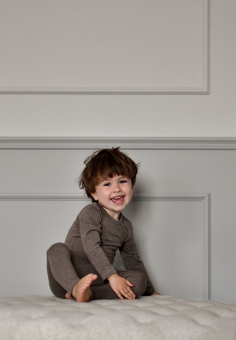 Vaikiškas ilgomis rankovėmis rudų spalvų smėlinukas su apvalia apykakle, dėvimas vaiko, sėdinčio ant šviesios, dygsniuotos audinio paviršiaus, besišypsančio ir žaidžiančio.