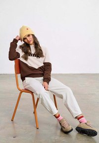 Mikina s blokem barev v hnědé a krémové barvě s textem, bílé kalhoty, béžové turistické boty a žlutá žebrovaná čepice s růžovým detailem.