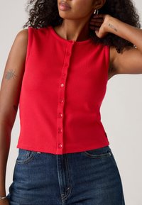 Haut sans manches rouge à côtes avec un col rond et une fermeture à boutons. Présente un design ajusté et une texture lisse, associé à un jean en denim foncé.