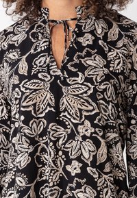 Blusa nera con un delicato motivo floreale beige, scollo a V con chiusura a fiocco e vestibilità comoda con maniche lunghe.