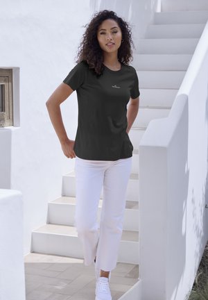 Schwarzes T-Shirt mit kurzen Ärmeln und Rundhalsauschnitt sowie Logo. Kombiniert mit weißen Hosen und weißen Sneakers, stehend auf hellen Treppen.