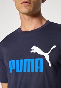 Navy blue t-shirt featuring a large white PUMA logo and text in bold blue. Crew neck design, made from smooth cotton fabric.