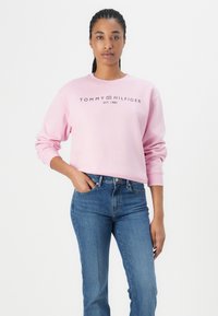 Rožinė džemperis su raštuotomis rankogaliais ir apykakle, puošiamas tamsiomis raidėmis užrašytu "Tommy Hilfiger" logotipu, derinamas su mėlynais džinsais.