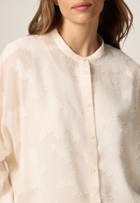Blusa button-up di crema chiaro con un motivo testurizzato, dotata di colletto alla cinese e sottili accenti bianchi. Tessuto morbido e trasparente.