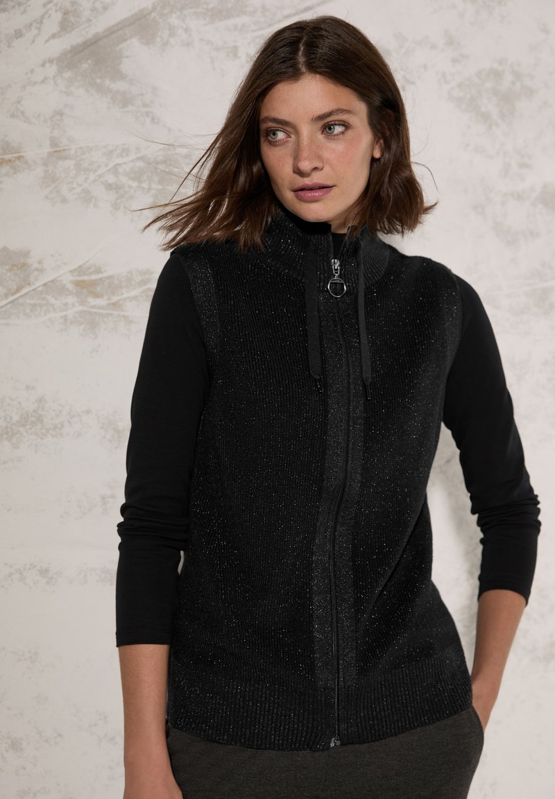 Gilet sans manches noir avec une fermeture éclair à l'avant, motif tricot texturé, col montant et accents brillants, superposé sur un haut noir à manches longues.