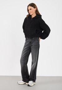Svart, strukturert fleece zip-up hettegenser med hette og frontlommer, kombinert med høyt skårede, lett slitte grå jeans og lyse joggesko.