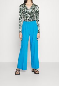 Langärmliges Oberteil in Schwarz und grünem Druck mit V-Ausschnitt, kombiniert mit hochgeschnittenen, weiten blauen Hosen und schwarzen Sandalen mit Riemchen.