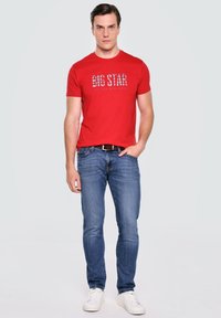 Červené bavlněné tričko s modrou grafikou "BIG STAR", kombinované s modrými džínami a bílými teniskami. Model stojí s uvolněnýma rukama.