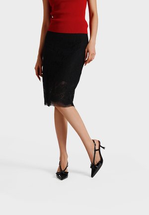 Mujer con una falda ajustada de encaje negro, top rojo sin mangas y zapatos de tacón altos negros puntiagudos con tiras en el tobillo, de pie sobre fondo blanco.
