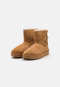 Tan suède enkellaarsjes met een zachte bontvoering, ronde neus en vetersluiting aan de zijkant, met gouden hardware accenten.