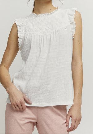 Kobieta ubrana w bezrękawnikową białą teksturowaną bluzkę z falbanami na ramionach i jasnoróżowe spodnie, stojąca z jedną ręką dotykającą spodni.